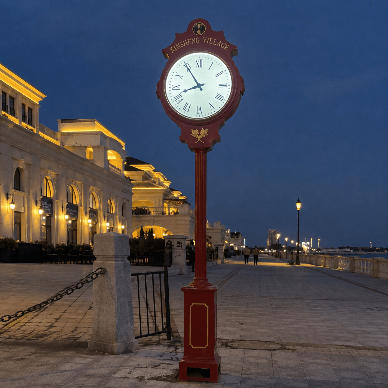 Illuminated red street clock along a waterfront promenade.