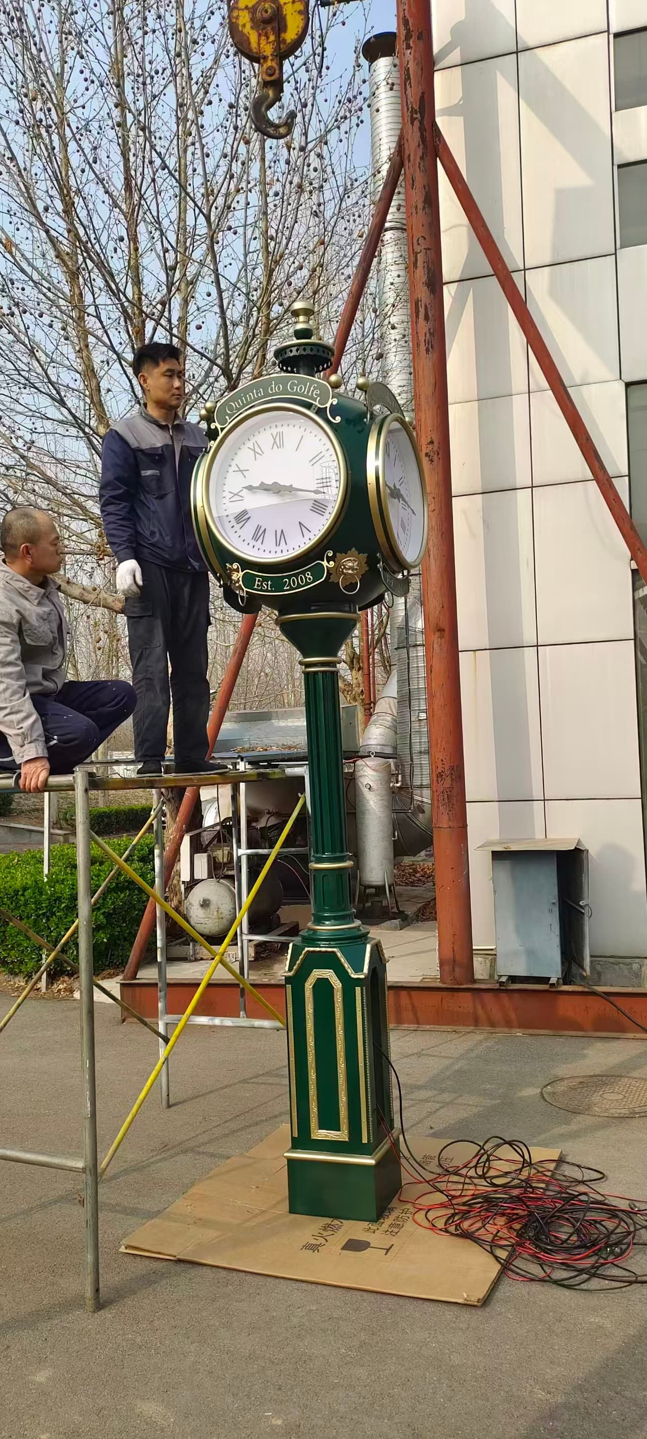 Two-sided green landscape clock being installed beside a building.